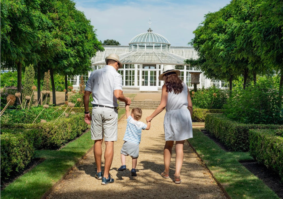 A family of three, dressed in light summer clothing, walks hand in hand toward a large glass conservatory surrounded by lush greenery on a sunny day.